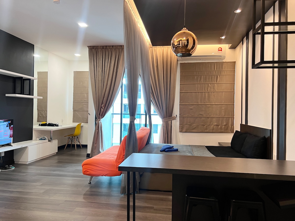 A studio unit features a spacious layout with large windows allowing natural light. An orange sofa contrasts with a black bed and dining area. Dark curtains frame the windows, while wall-mounted shelving and a television enhance convenience.