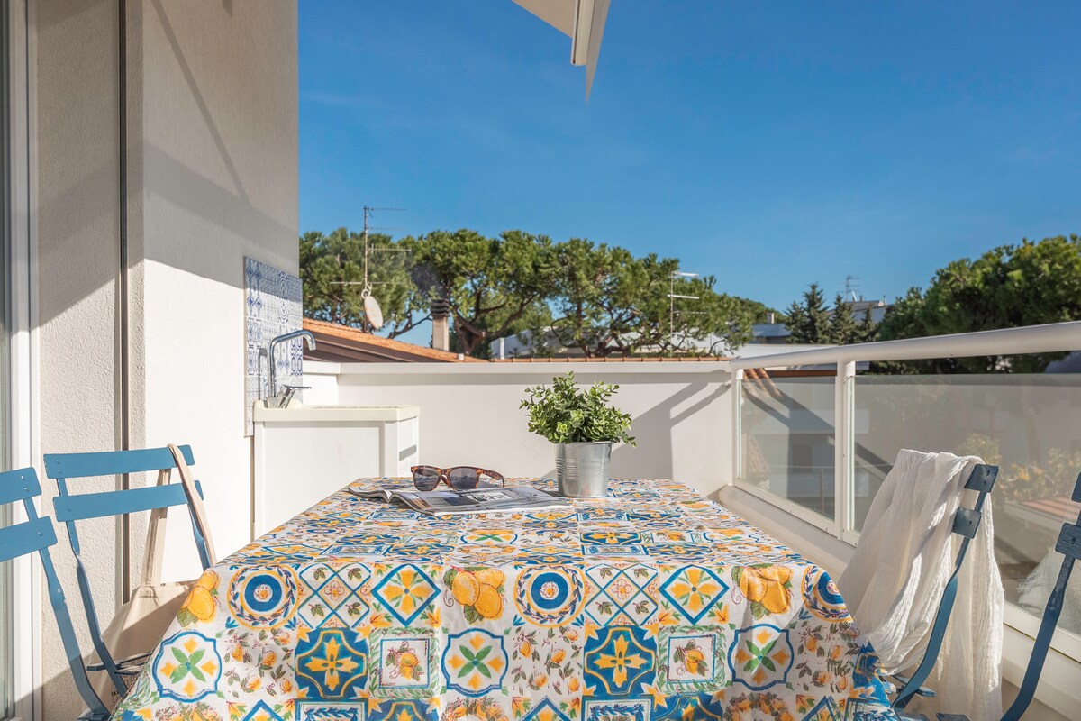 A balcony features a table covered with a decorative cloth, complemented by two blue chairs. A small potted plant sits at the center of the table, with sunglasses resting nearby. The view includes clear skies and greenery in the background, enhancing the outdoor setting.