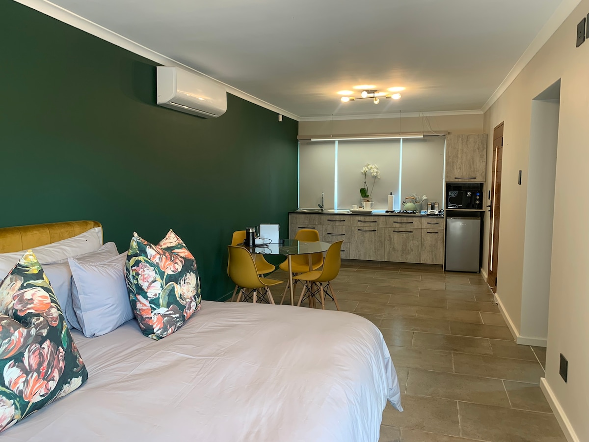 A spacious sleeping area features a King bed adorned with decorative pillows. A kitchenette is visible in the background, equipped with modern appliances. A small dining table with four chairs is positioned nearby, contributing to the open-plan layout. Light filters in through large windows.