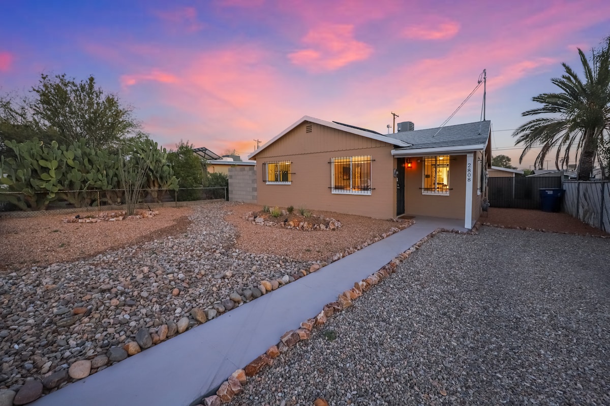 The exterior of the cozy home features light-colored walls and a welcoming front path lined with pebbles. Surrounding landscaping includes sparse greenery and decorative rocks, with a spacious yard visible. A pink and purple sky enhances the inviting atmosphere at sunset.