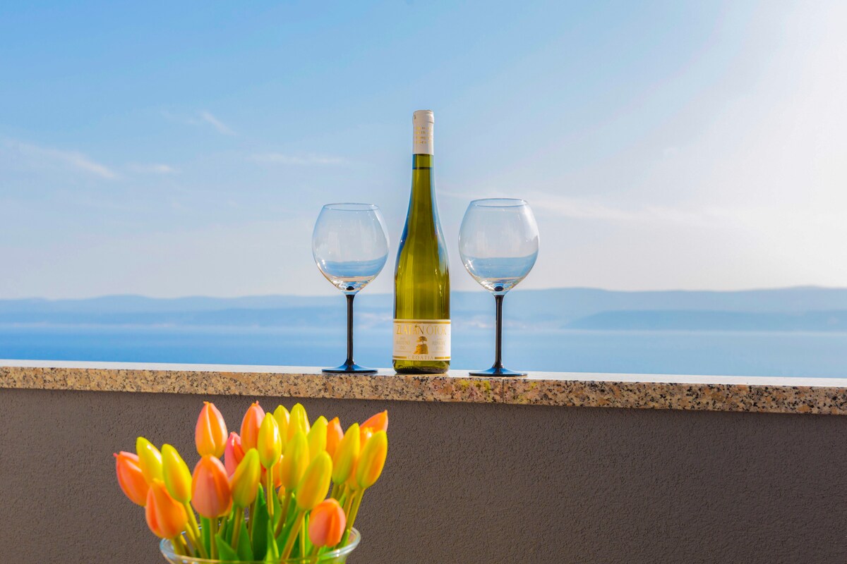 A scenic view from a terrace is highlighted by a bottle of wine and two elegant glasses perched on a stone surface. In the foreground, a vibrant bouquet of tulips showcases a mix of yellow and orange flowers, adding a touch of color against the backdrop of calm waters.