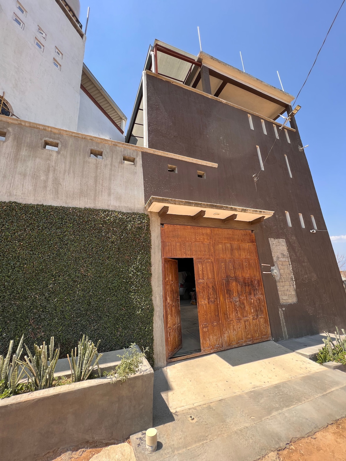 A modern exterior of a building is highlighted, showcasing a distinctive entrance with a large wooden door. The facade features a combination of earthy tones and textures, complemented by greenery along the walls. The clear blue sky provides a bright backdrop.