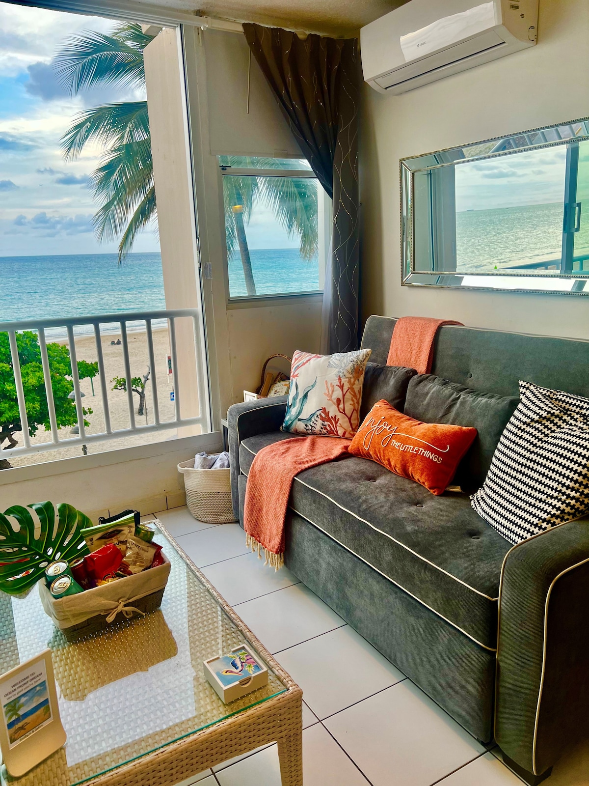 A comfortable living area features a queen-size sofa bed, adorned with decorative pillows and a throw blanket. A large window offers views of the beach and ocean, with natural light illuminating the space. A small table with refreshments is also visible.