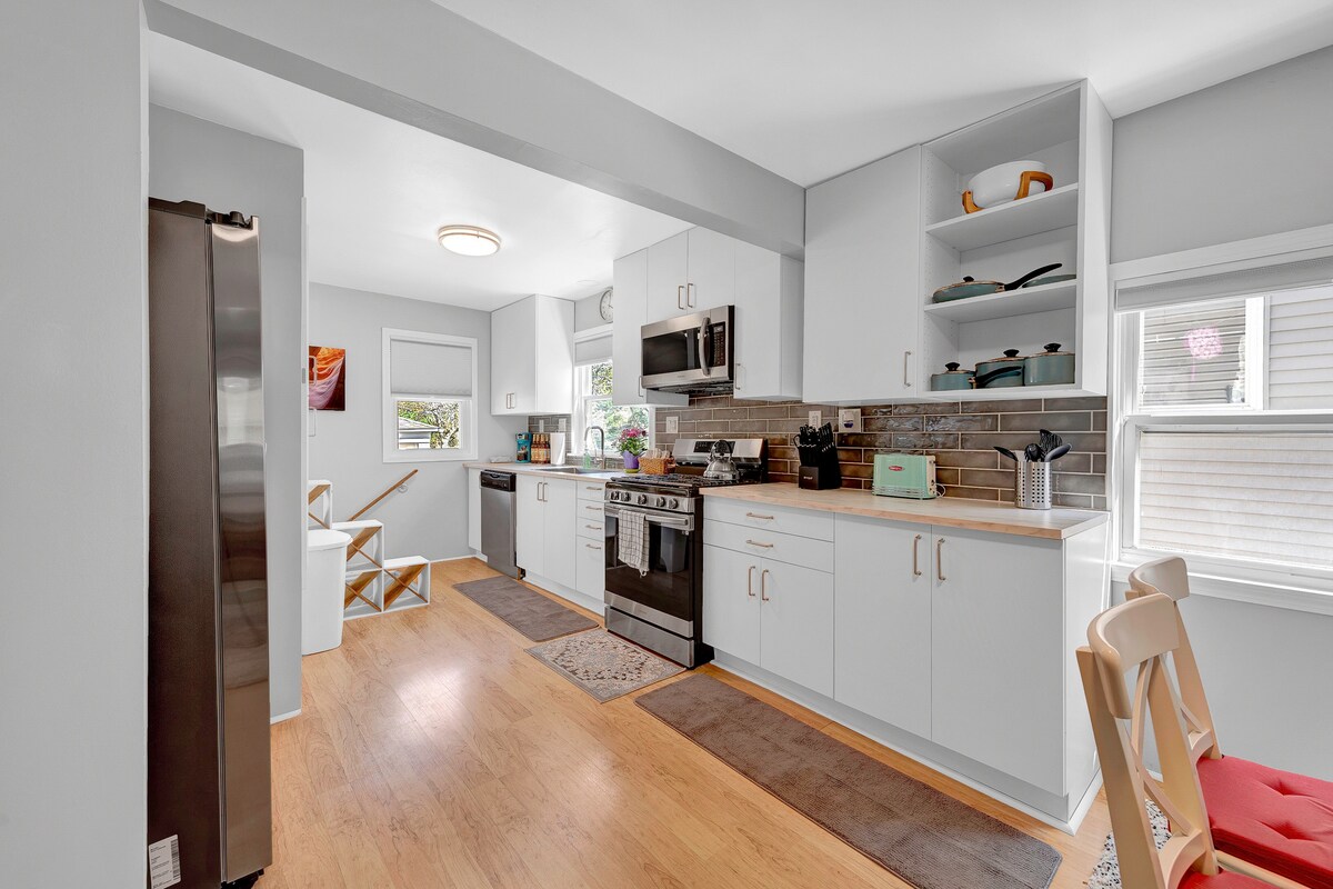 A modern kitchen features stainless steel appliances and white cabinetry, with a neutral countertop. Natural light filters through a nearby window. Open shelves display colorful kitchenware, while a dining area with a wooden table is visible in the background, creating a spacious and functional layout.