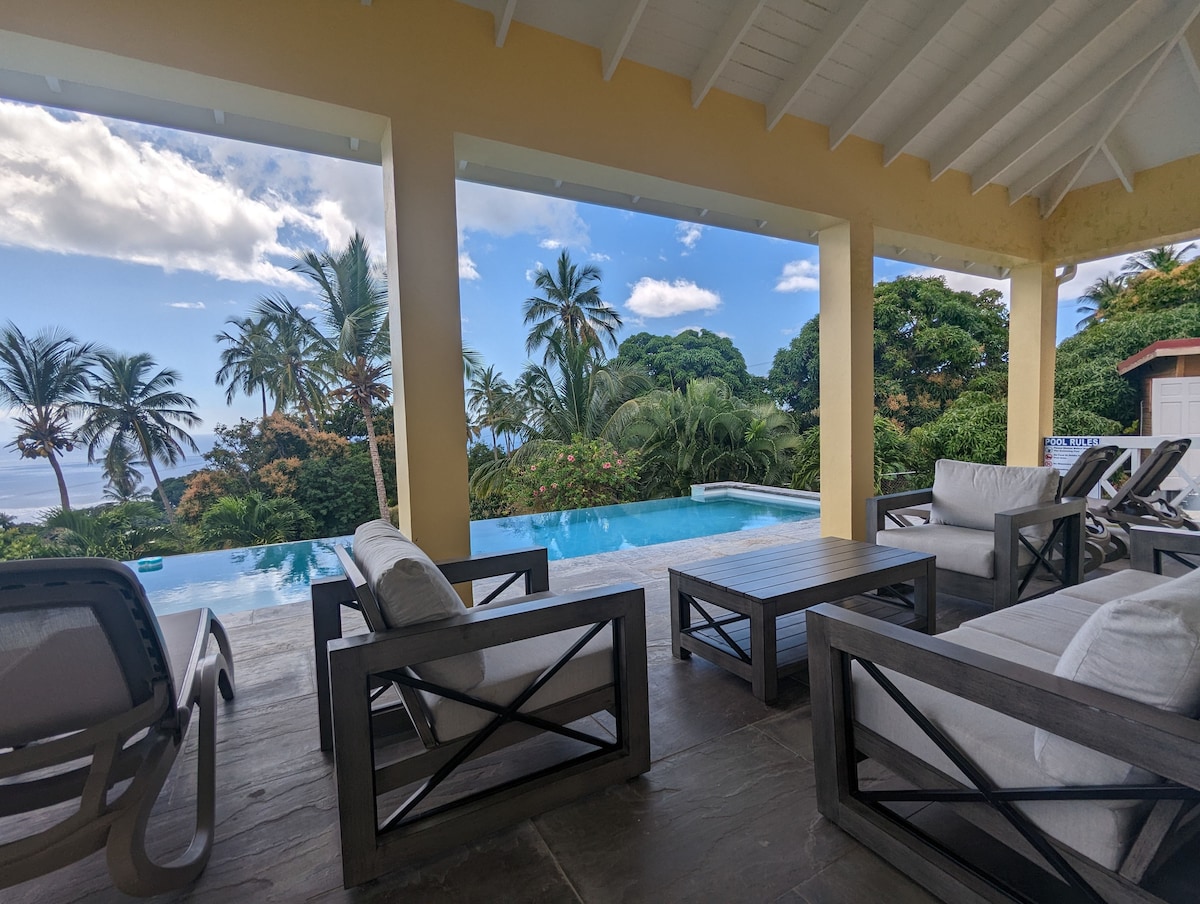 An outdoor lounge area features comfortable seating arranged around a wooden table, overlooking a serene swimming pool. Lush greenery and palm trees frame the view, while a bright blue sky adds to the tranquil atmosphere.