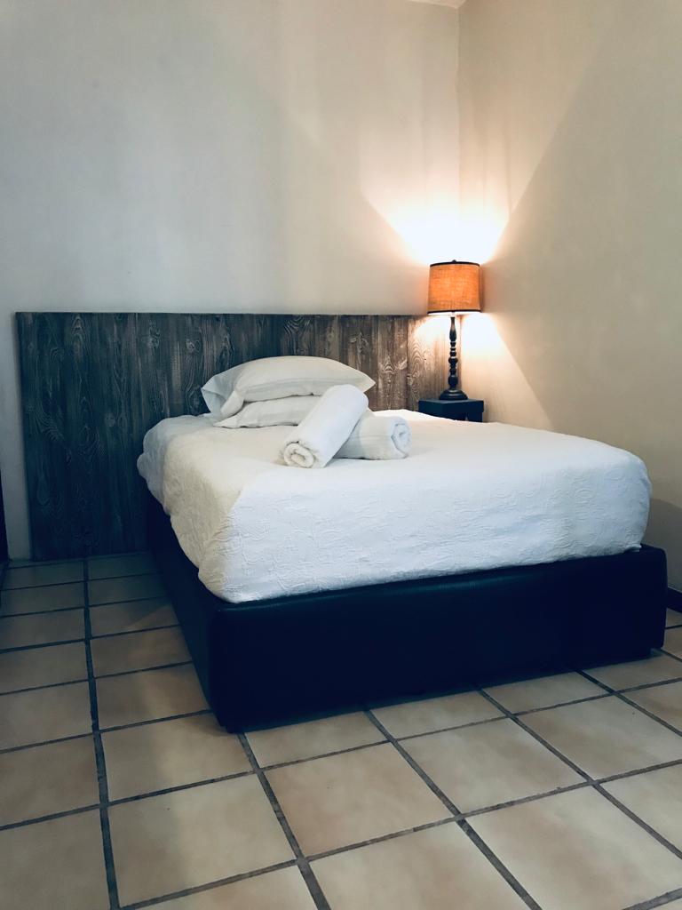 A cozy sleeping area features a spacious bed with white bedding and rolled towels resting on top. A bedside lamp provides soft lighting, while the warm tone of the walls complements the light-colored tiled floor.