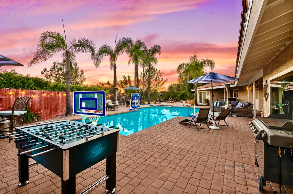 The outdoor area showcases a sparkling pool surrounded by lounge chairs and umbrellas. A foosball table is positioned in the forecourt, with palm trees lining the background. The scene is enhanced by a colorful sunset sky, creating a relaxing atmosphere for guests.