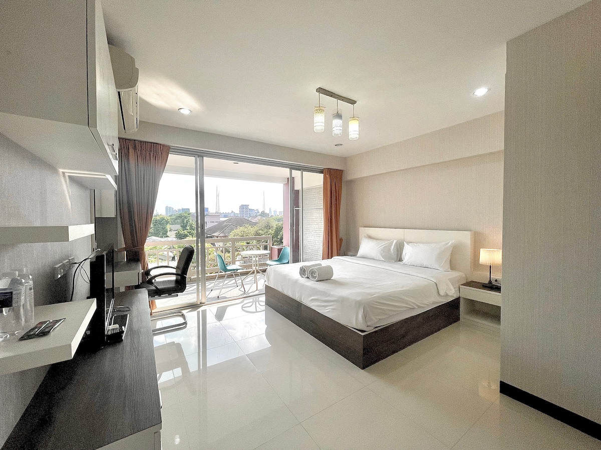 A bright studio features a spacious bed positioned against a light-colored wall. Large glass doors open to a balcony with city views. A compact kitchen area includes a coffee maker and cooking space, complemented by modern lighting fixtures throughout the room.