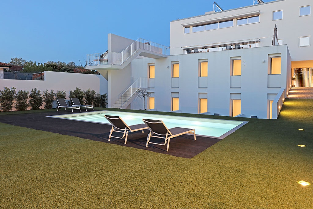 An outdoor pool area is surrounded by lush green grass, featuring a spacious pool illuminated in soft lighting. Comfortable lounge chairs are positioned near the water, and a white building with large windows stands in the background, creating a serene environment.