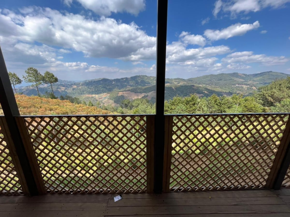 A scenic view from a covered balcony reveals rolling hills and lush greenery, under a partly cloudy sky. The wooden railing features a lattice design, complementing the natural surroundings and providing a serene space to enjoy the outdoors.