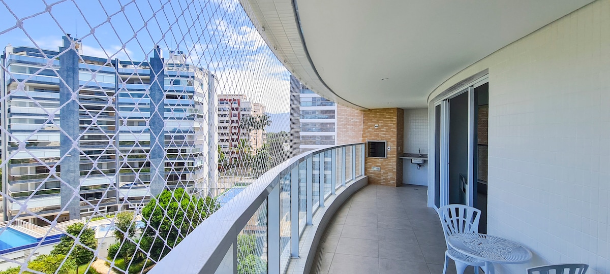 A wide balcony features a netted safety barrier, with a view of neighboring buildings and greenery. A small table and a chair are positioned for comfortable seating. The space is designed for relaxation, complemented by bright natural light and an open atmosphere.