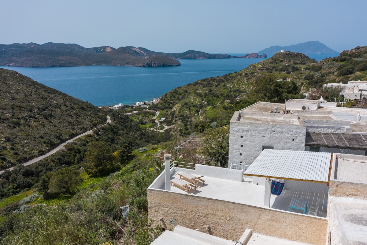 An expansive view reveals rolling green hills leading to a serene blue sea, with the coastline and distant mountains in the background. A private terrace, equipped with outdoor furniture, offers a clear perspective of the tranquil landscape and nearby village, inviting relaxation.
