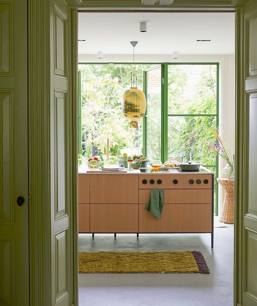 A modern kitchen is viewed through an open doorway, showcasing warm wooden cabinetry and a central island with cooking facilities. Large windows allow natural light to flood the space, surrounded by greenery. Decorative items are arranged on the counter, enhancing the inviting atmosphere.
