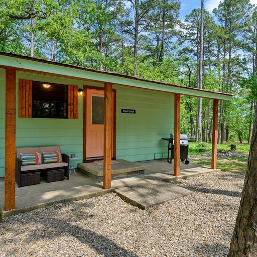 The exterior of the cabin is shown, featuring a front porch with a seating area and a designated grilling spot. Surrounded by tall trees, a light green exterior complements the natural setting. The entry door stands out with a coral hue against the wooden frame.