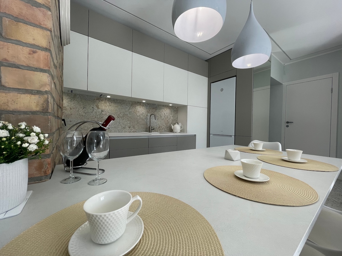 A modern kitchen featuring sleek cabinetry in grey and white tones. A spacious countertop is set with placemats, cups, and wine glasses. Soft pendant lighting hangs above, illuminating the glassware and a decorative plant nearby.