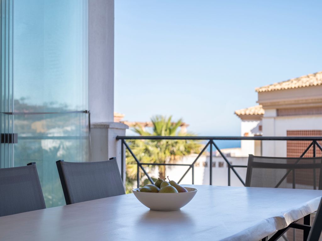 An outdoor dining area features a large table with minimalist chairs. A bowl of fresh fruits is positioned at the center. In the background, palm trees and distant ocean views are visible, complemented by a clear blue sky.