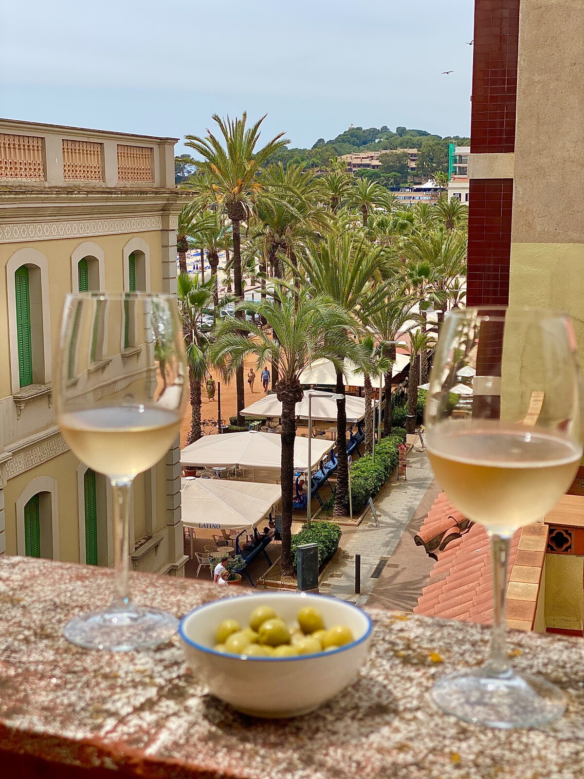 A balcony view features two glasses of white wine and a small bowl of olives, with palm trees lining the promenade below. Sunlight filters through the tropical scene, revealing outdoor seating areas at restaurants and cafes, creating a lively coastal atmosphere.