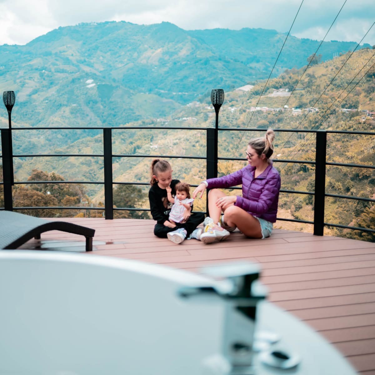A seating area is positioned on a wooden deck, overlooking rolling mountains in the background. Two individuals sit comfortably on the surface, surrounded by a serene landscape. A modern bathtub with a faucet is visible in the foreground, adding to the tranquil setting.