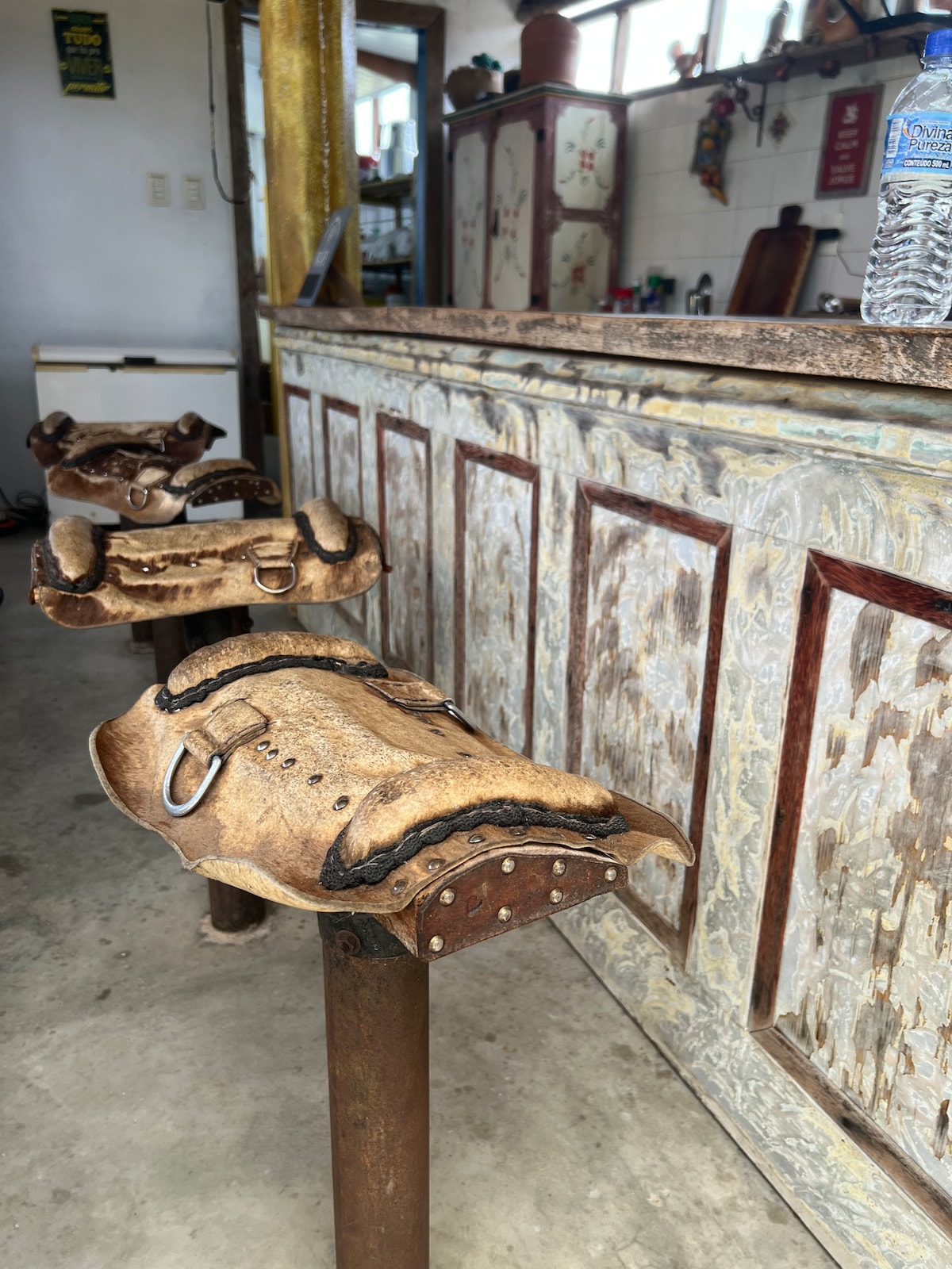 Three uniquely designed bar stools, crafted from leather and metal, are positioned in front of a rustic bar. The bar features a weathered finish with decorative paneling and a variety of personal items visible on the shelves behind it.
