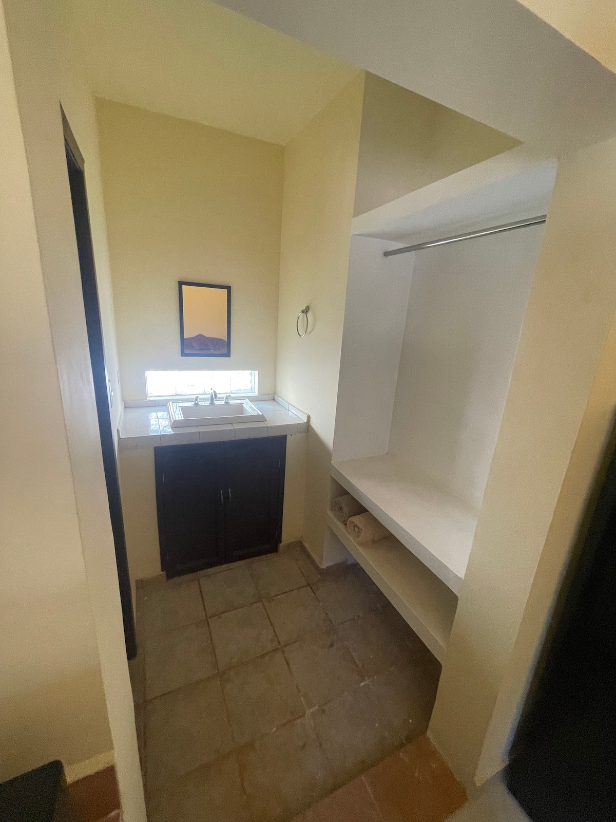 The bathroom area features a compact sink with a mirror above, accompanied by a small window allowing natural light. A closet space and shelving are present, providing practical storage options. Neutral-colored walls and tiled flooring create a simple, clean aesthetic.