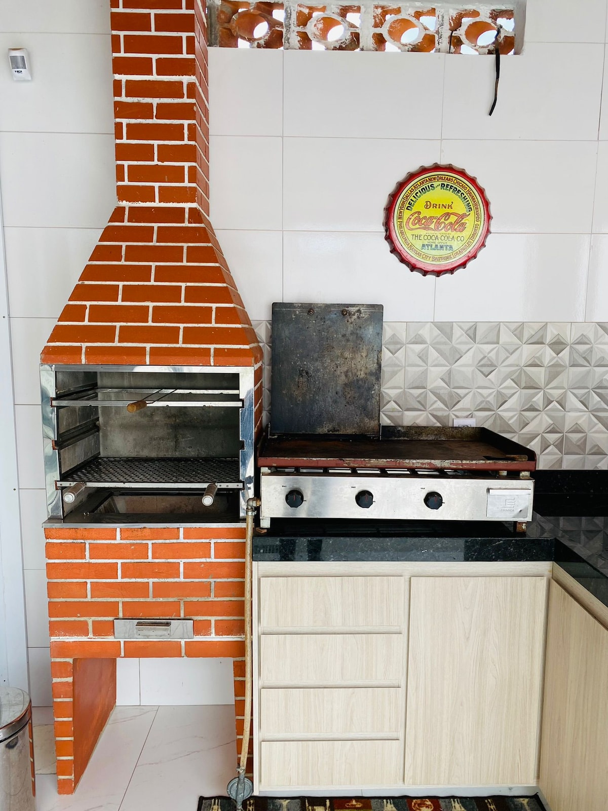 The outdoor kitchen features a brick barbecue grill with a metal griddle adjacent to it. Light-colored cabinetry provides storage beneath a dark countertop, while decorative wall tiles offer visual interest. A round sign hangs above, adding a touch of color.