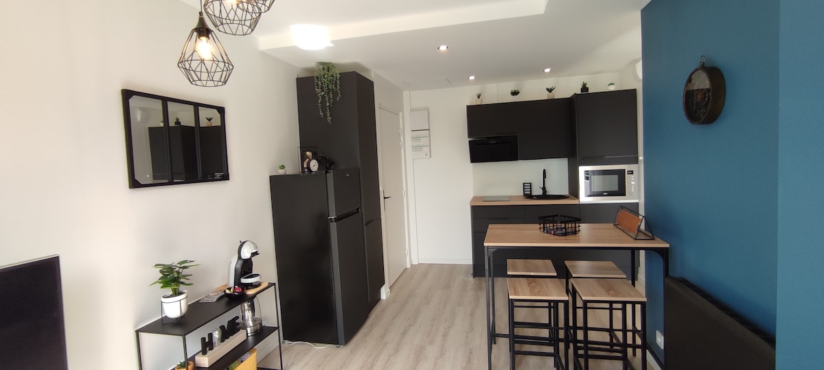The modern kitchen area is equipped with sleek black cabinetry and stainless steel appliances, including a microwave and an upright refrigerator. A small dining table with two stools offers a cozy space for meals. The adjacent living area features a simple shelving unit with plants.