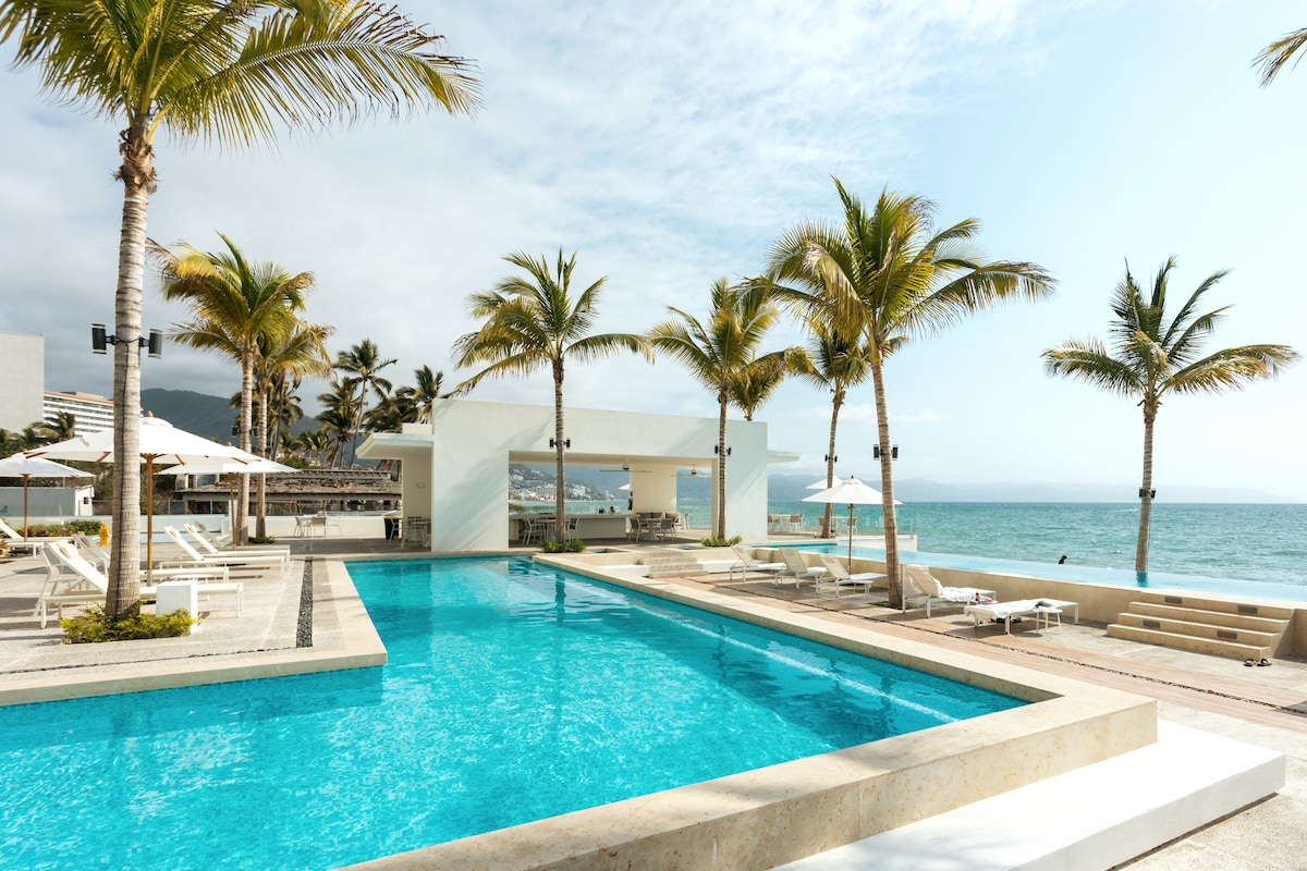 The image captures a tranquil pool area surrounded by palm trees. A spacious pool reflects blue skies and is complemented by lounge chairs and umbrellas. The ocean is visible in the background, providing a serene seaside atmosphere.