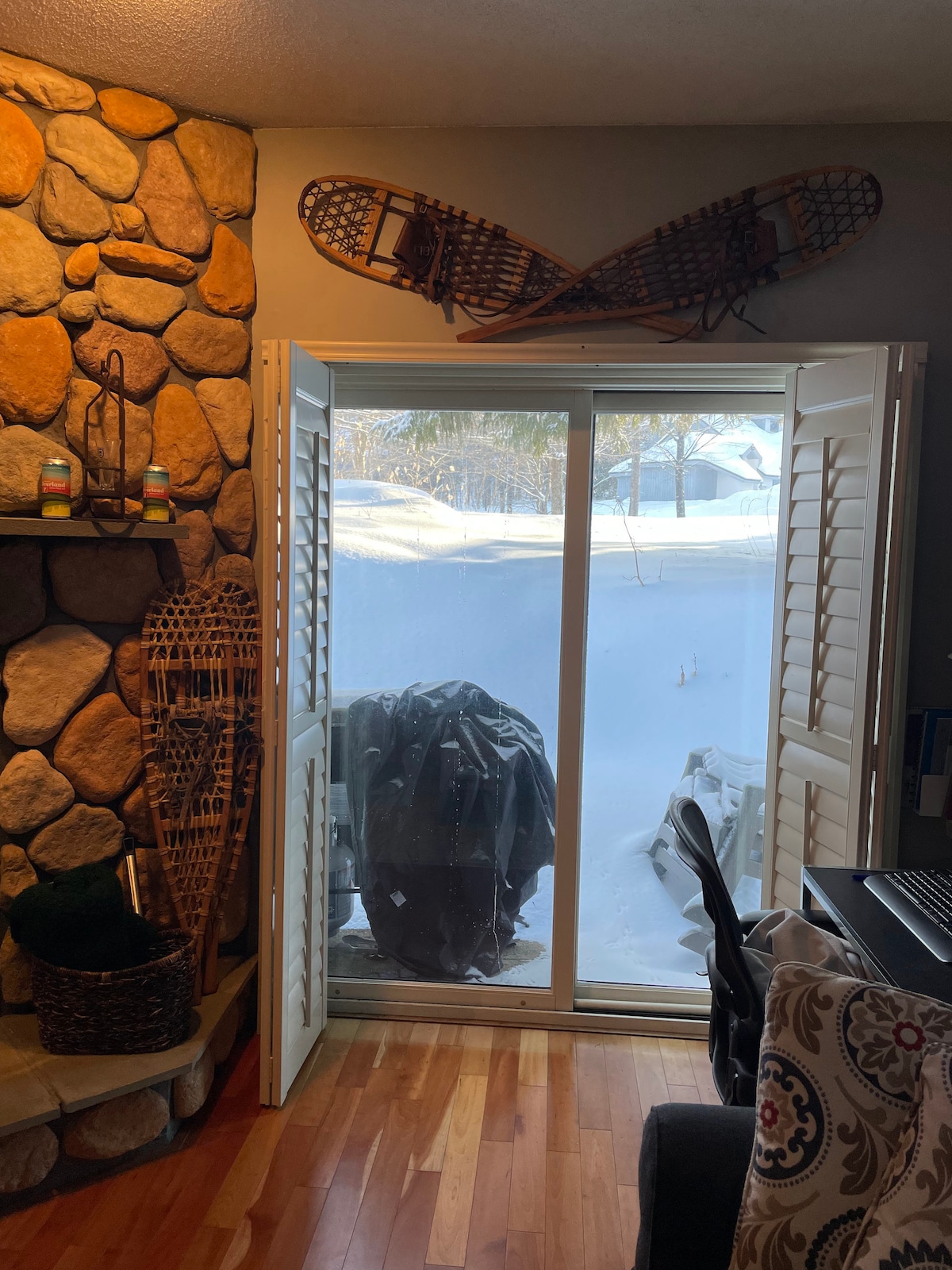 A sliding glass door with white shutters is framed by a stone wall, revealing a snowy outdoor landscape. Snowshoes are mounted above the door, adding a ski lodge element. A dark covered grill is positioned outside, while a cozy workspace is visible to the right.