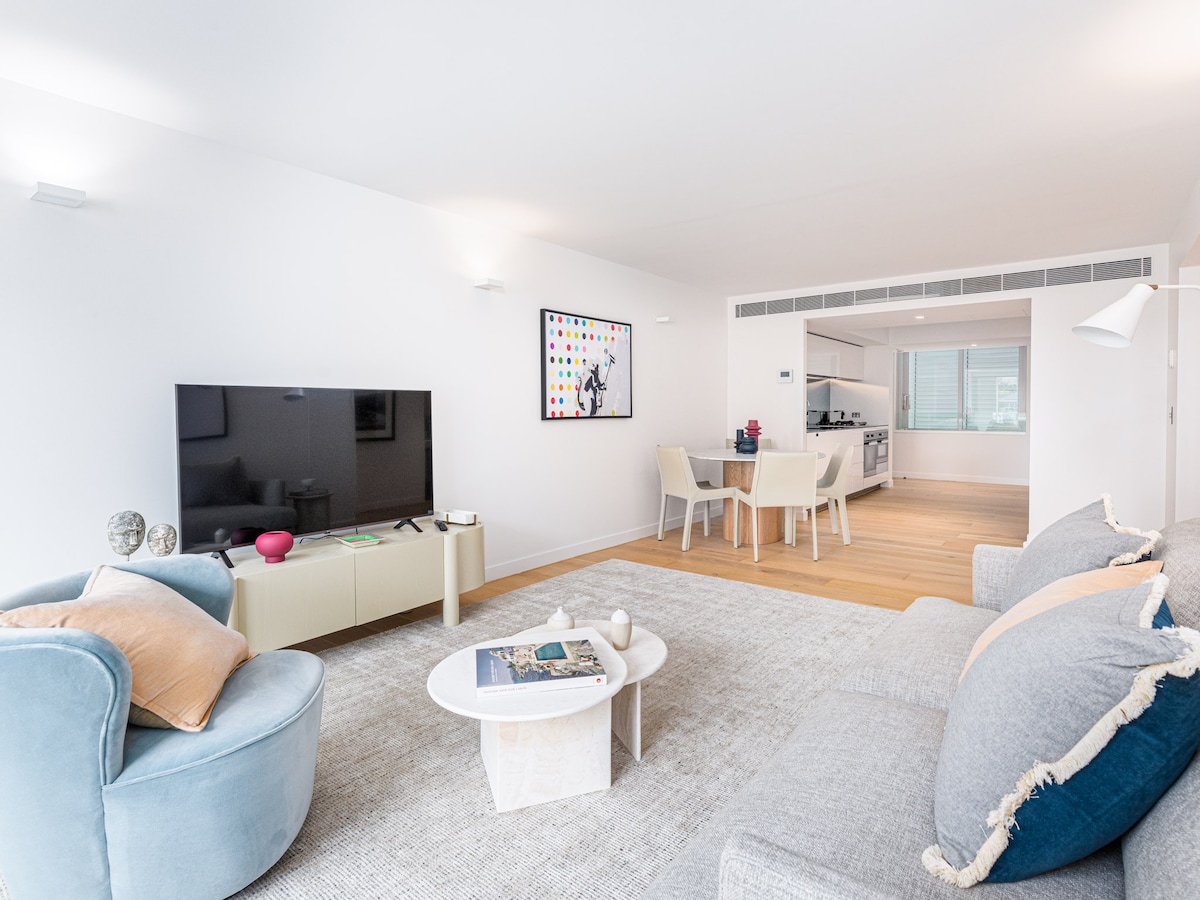 A modern living area features a comfortable gray sofa and a round coffee table, topped with a book. A soft blue armchair is positioned nearby. Natural light fills the space from large windows, and a dining area is visible in the background with a table and chairs.