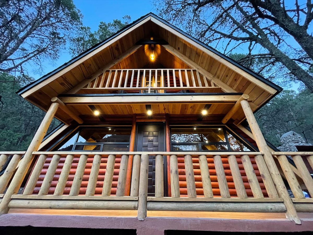 The cabin's architectural features are highlighted, showcasing a sloped roof constructed of wood. A spacious balcony is framed by log railings, while large windows allow natural light to flood the interior. Surrounding trees enhance the serene setting.