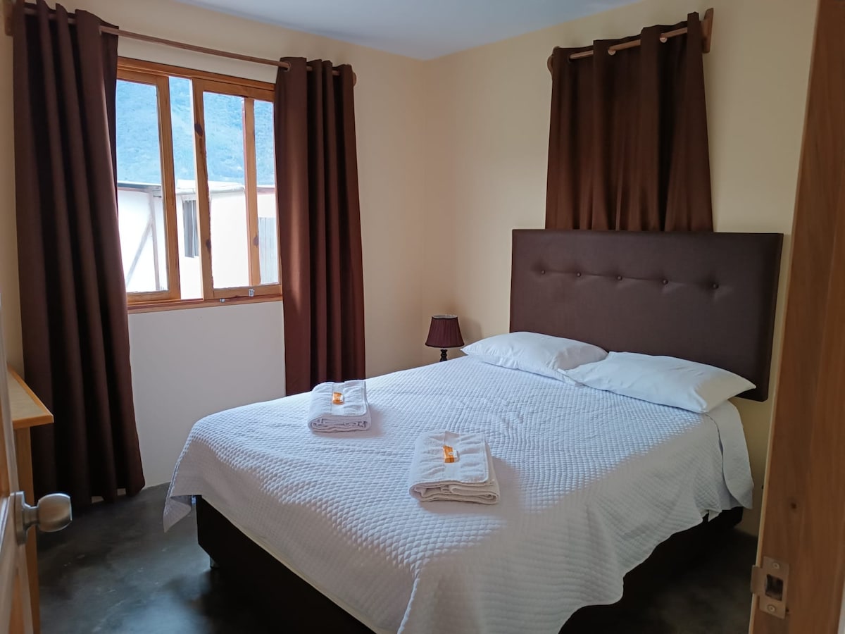 A cozy bedroom features a spacious bed with white linens, neatly arranged towels, and a bedside lamp. Natural light enters through a large window, bordered by dark curtains. A wooden dresser is positioned nearby, contributing to the room's inviting ambiance.