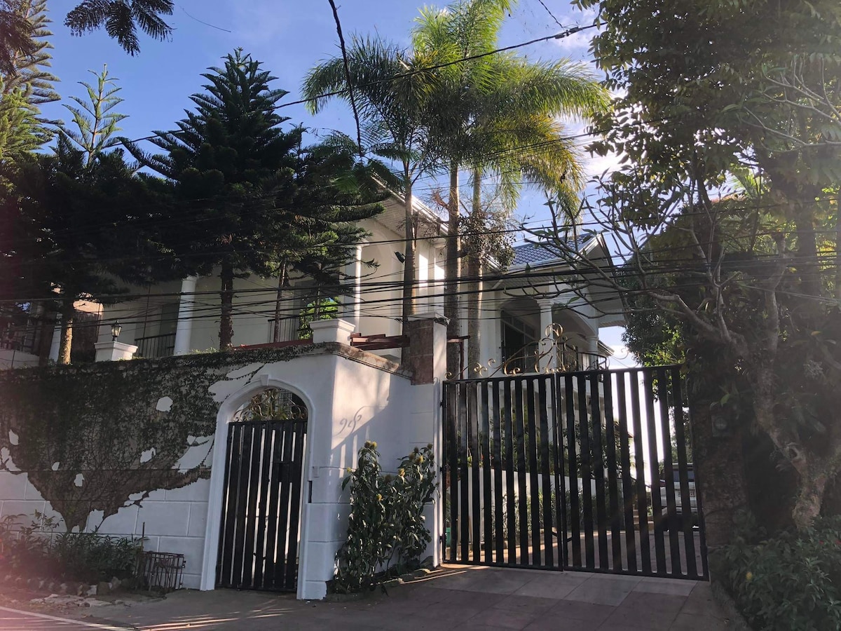A secure gated entrance with vertical black slats is framed by lush green landscaping. Towering palm and pine trees are visible, alongside a white structure that features a second level, hinting at the spaciousness of the property. Bright blue sky peeks through the foliage.