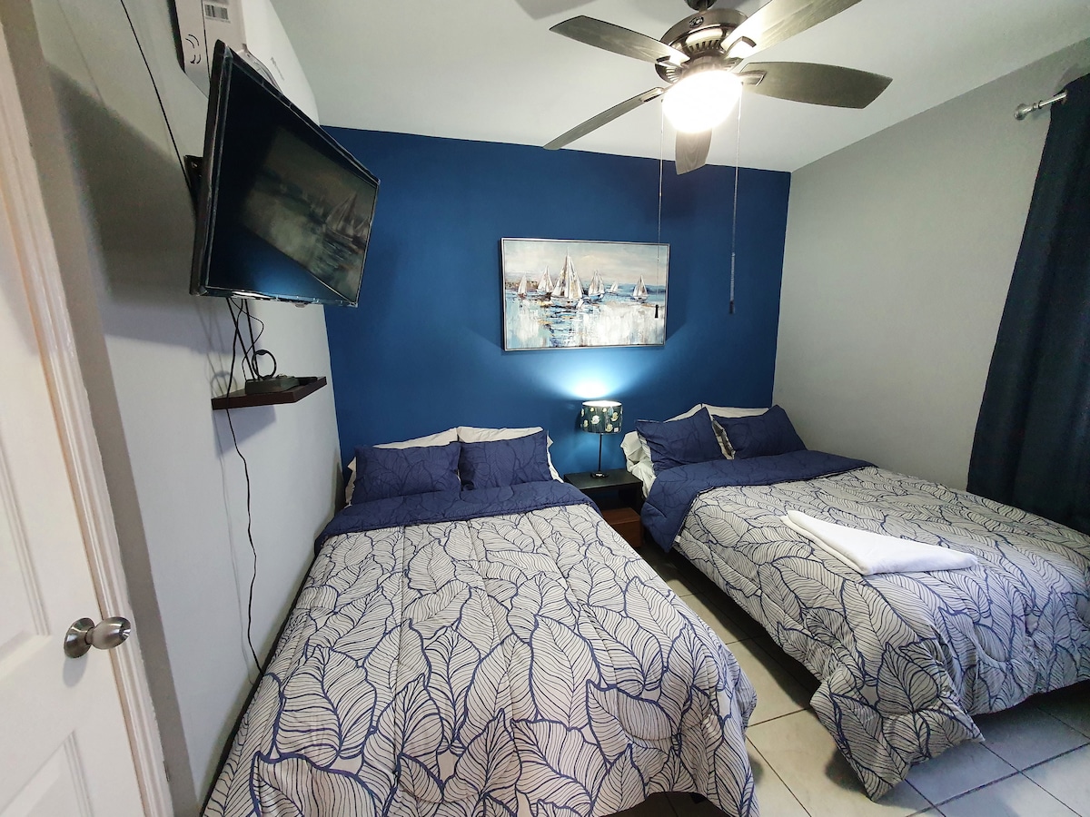 A private room features two spacious beds adorned with patterned linens. A flat-screen television is mounted on the wall opposite the beds, and a piece of nautical-themed artwork adds character. Natural light enters through a window, complemented by dark curtains.