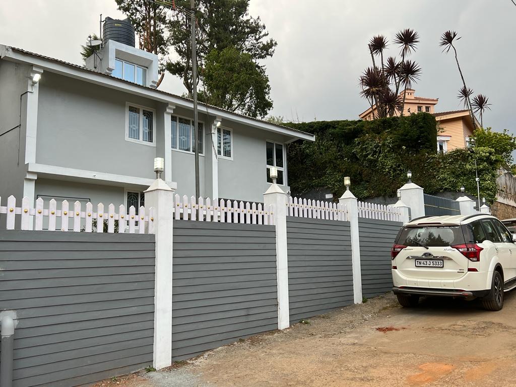A two-story house is shown with a light grey exterior, framed by a white picket fence. The manicured surroundings include green bushes and trees. A parked white SUV is visible in the driveway, with a gravel surface leading to the entrance.
