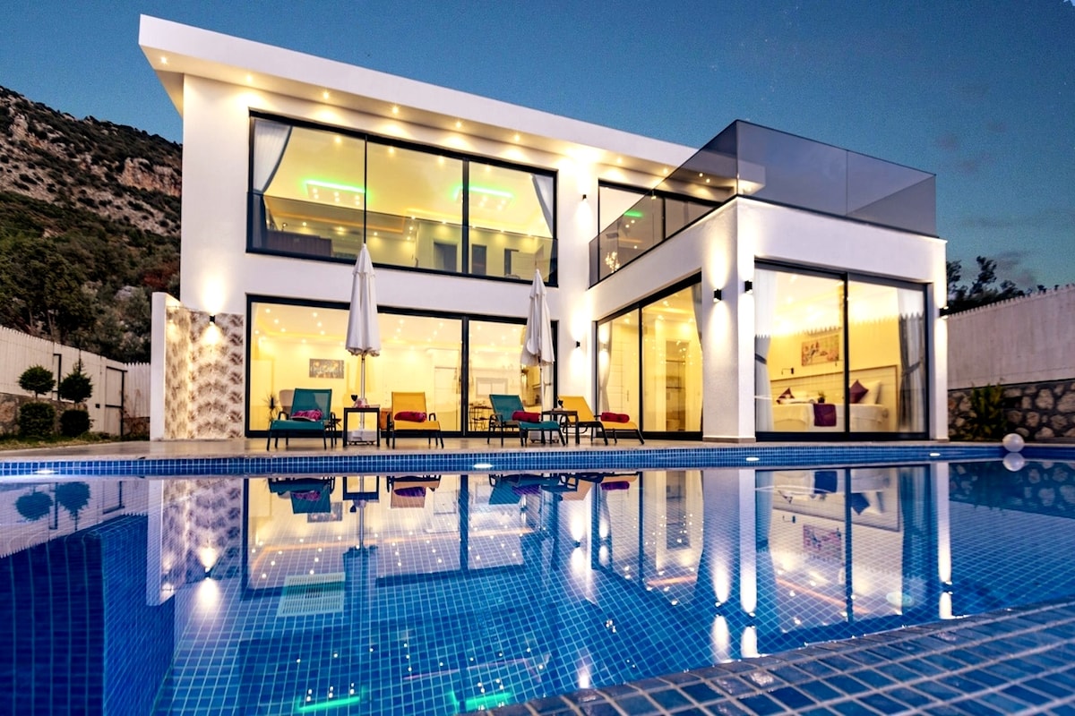 An elegant villa is reflected in the clear waters of an infinity pool, surrounded by lounge chairs. The modern architecture features expansive glass walls and a spacious terrace, allowing for abundant natural light. Umbrellas offer shade, enhancing the inviting outdoor space.