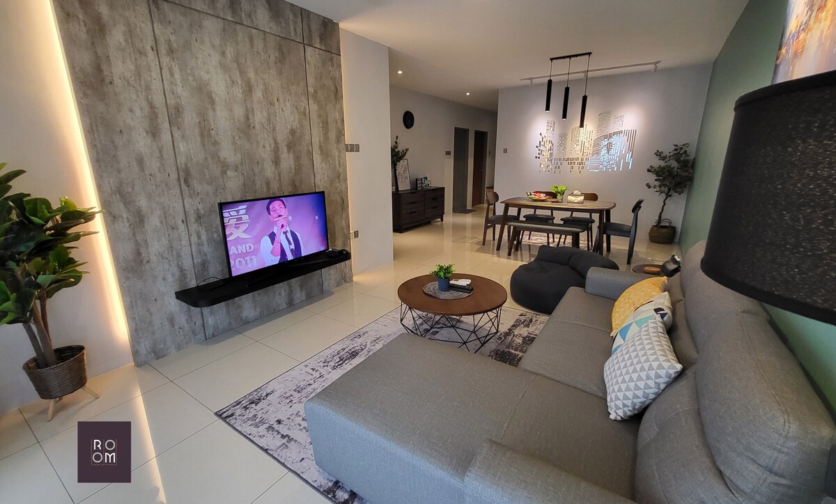 A modern living area features a comfortable grey sofa with decorative cushions. A wall-mounted television displays content, while a wooden dining table is set nearby. Plants are positioned in corners, adding a touch of greenery to the space. Soft lighting creates a relaxed ambiance.