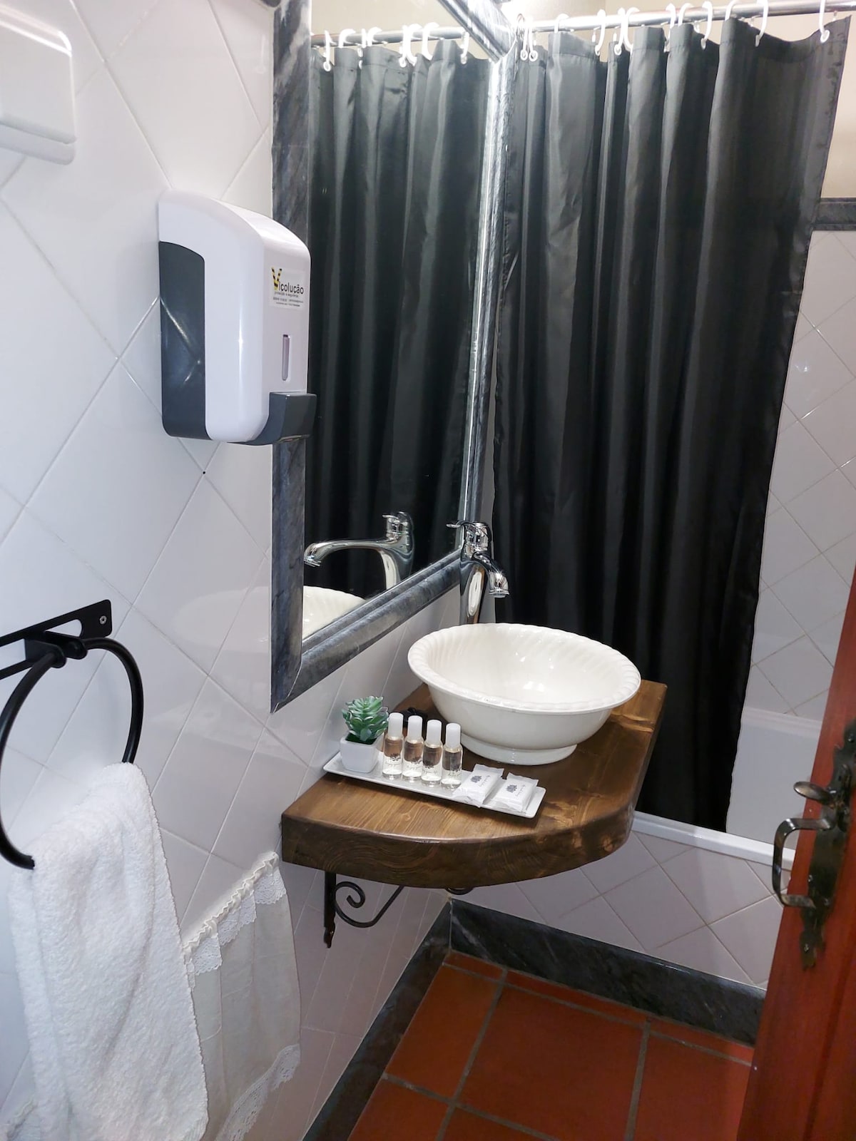 A compact bathroom features a wooden shelf supporting a round basin and an arranged set of toiletries. Black curtains frame the shower area, while a towel hangs neatly on a rack. A soap dispenser is mounted on the wall, complementing the overall design.