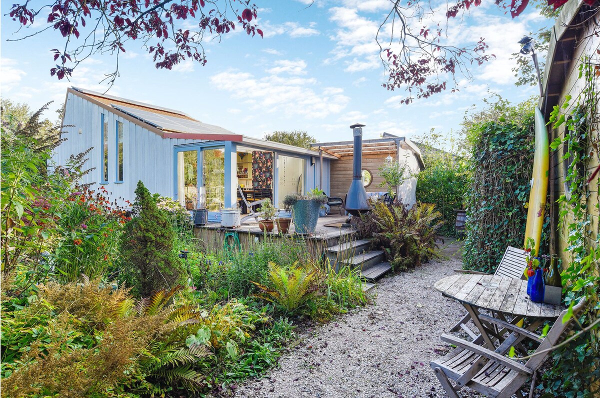 A charming outdoor area is framed by lush greenery and colorful plants. Steps lead to a cozy seating arrangement and a modern chimney, while large glass doors offer a glimpse into the inviting interior space. Natural elements create a serene ambiance.