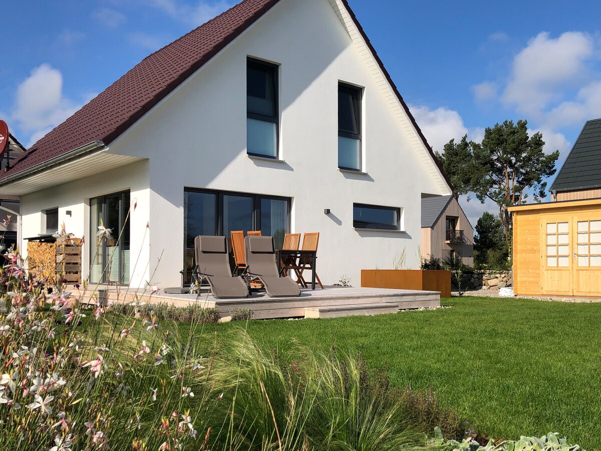 The white house features a sloped roof and large windows, allowing natural light to fill the interior. A spacious wooden terrace is furnished with two lounge chairs, while a well-maintained garden surrounds the structure, adding greenery and a serene atmosphere.