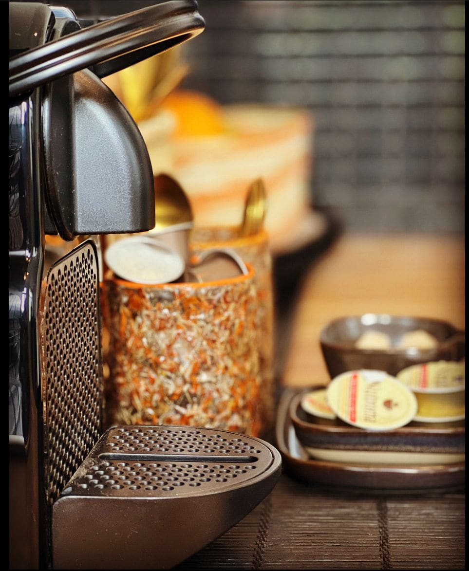 A coffee machine is positioned prominently with stylish mugs and assorted coffee capsules nearby. Various utensils are arranged, showcasing an organized setup for beverage preparation. The scene is complemented by a soft wooden surface and warm light, creating an inviting atmosphere for coffee enthusiasts.