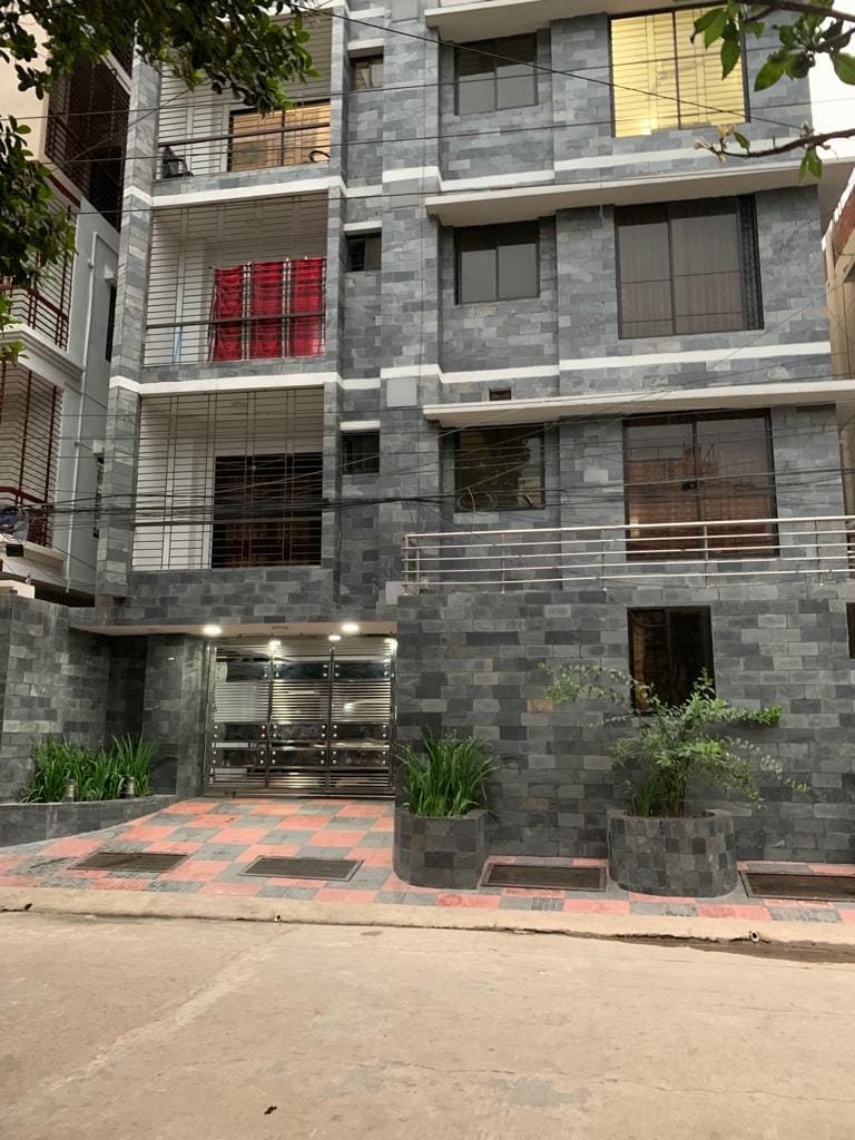 The exterior of the building is characterized by modern grey stone cladding, adorned with large windows and a contemporary design. A landscaped entrance features potted plants and an inviting pathway leading to the main entrance, which is glass-fronted and well-lit.