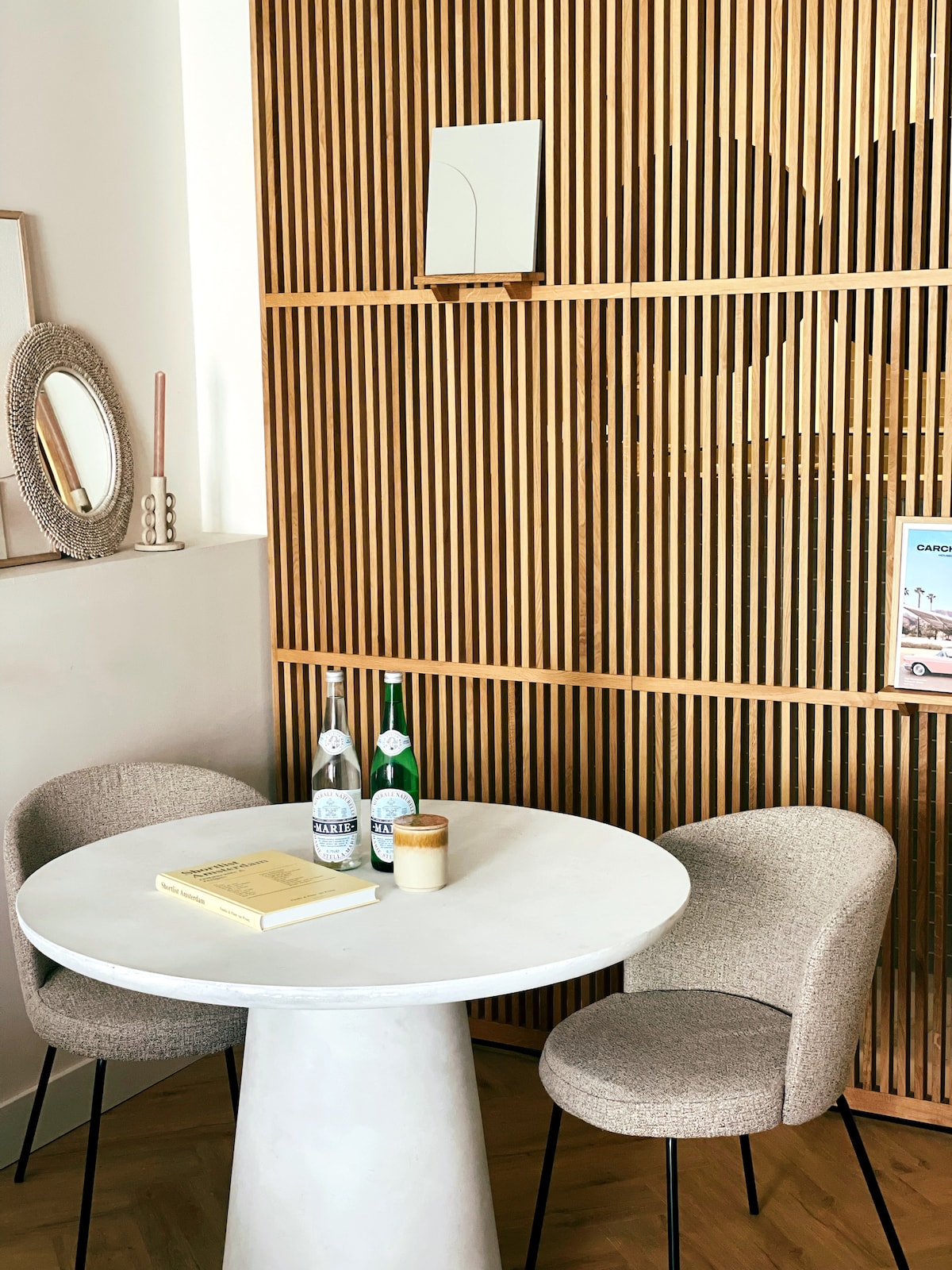 A round stone table is positioned in front of a wooden slatted wall. Two upholstered chairs are placed on either side of the table, which is adorned with two bottles of sparkling water and a small candle. Soft lighting enhances the minimalist decor.