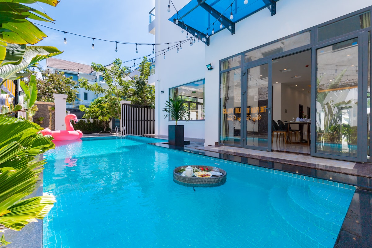 An outdoor swimming pool is surrounded by lush greenery, featuring a floating tray with fresh fruit. Colorful lights are strung overhead, and large glass doors provide a view into the adjacent living area. A pink float adds a playful touch to the serene setting.