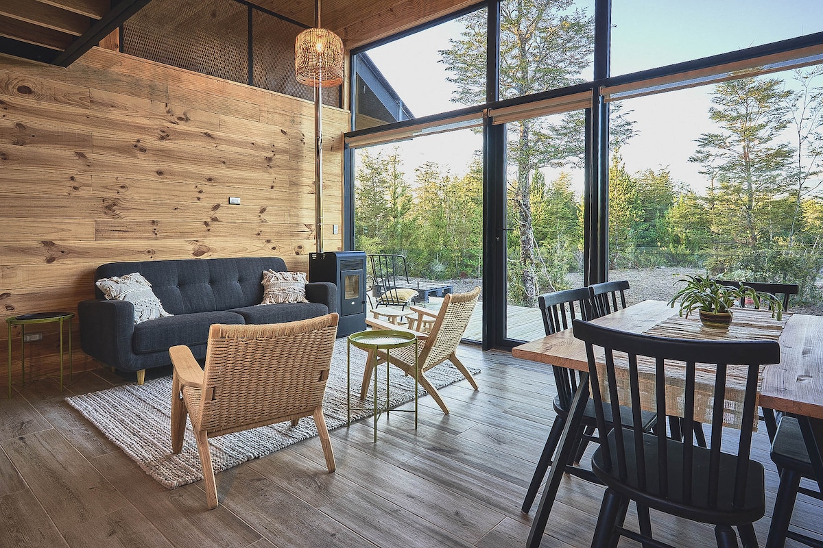 A cozy living area features natural wood walls and large glass doors that open to the outdoor scenery. Comfortable seating includes a dark sofa and woven chairs. A rustic wooden dining table sits alongside, allowing for family gatherings. Warm light filters through the windows.