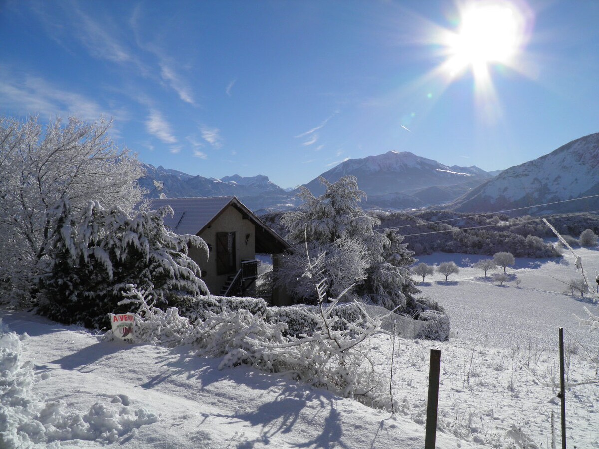 A snow-covered landscape showcases a charming house surrounded by frosted trees. Majestic mountains rise in the background under a bright sun, reflecting off the white snow. A clear blue sky enhances the serene winter scene.