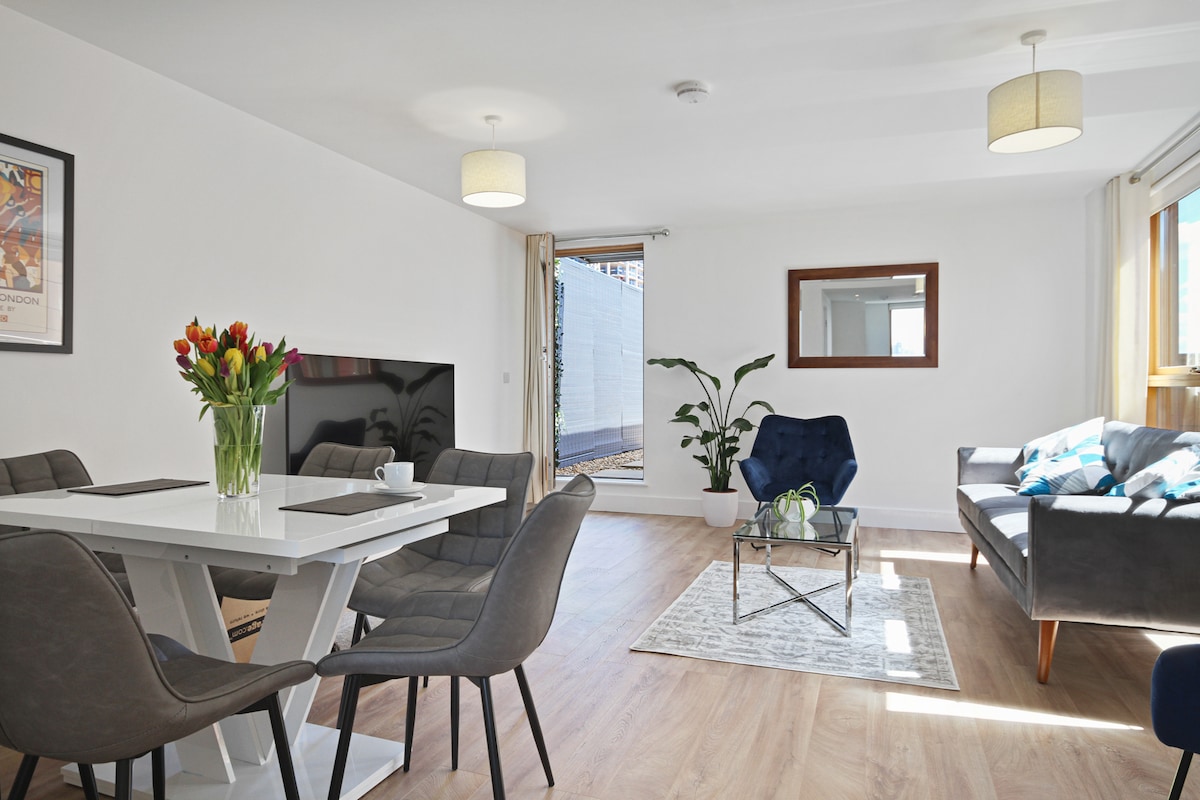 An open-plan living and dining area features a white dining table surrounded by six chairs, complemented by a comfortable blue armchair and a grey sofa. A large window allows natural light to illuminate the space. A vase of fresh flowers adds a touch of color.