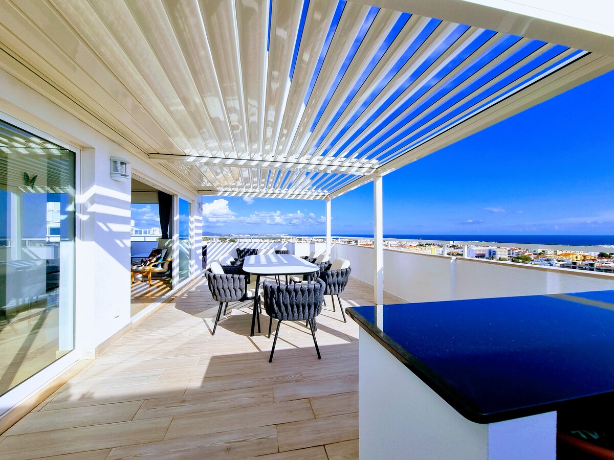 A spacious terrace is featured with a dining table surrounded by comfortable chairs, sheltered by a bioclimatic pergola. The stunning views of the sea and city extend beyond, providing a bright and inviting outdoor space under a clear blue sky.