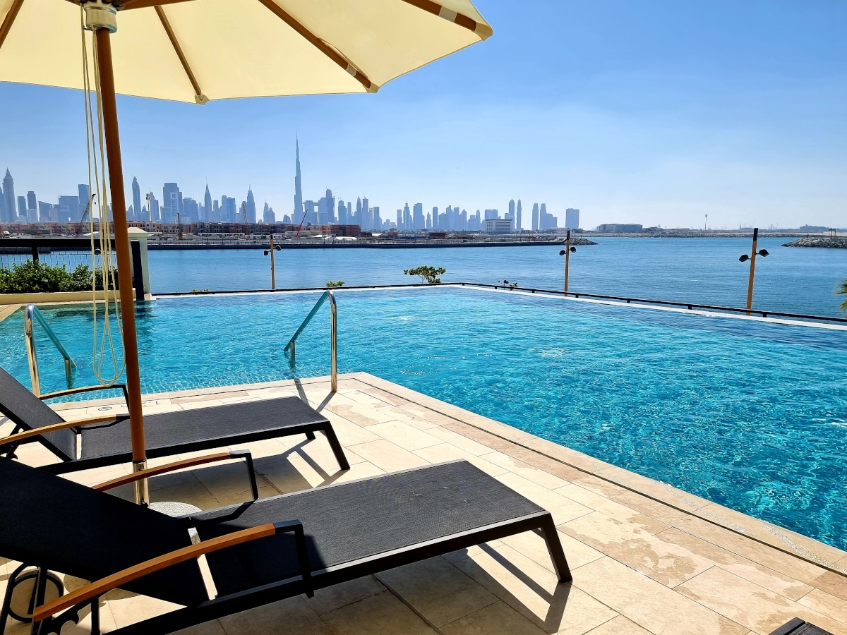 A serene pool area features two lounge chairs positioned under a large umbrella, overlooking a shimmering body of water. The city skyline, including the Burj Khalifa, provides a striking backdrop against a clear blue sky.