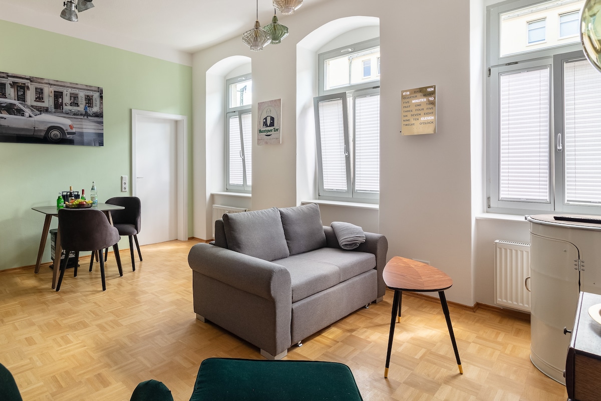 A bright living space features large windows allowing natural light to fill the room. A gray sofa is positioned next to a small wooden coffee table. In the background, a dining table is set for four, complemented by a modern green accent chair.