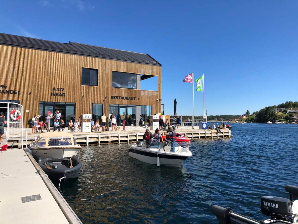 A modern wooden building houses a restaurant and shops along the waterfront. Patrons are seated on the deck, enjoying views of the water and boats. Multiple flags flutter in the breeze, and a few boats are docked nearby, adding to the lively atmosphere.